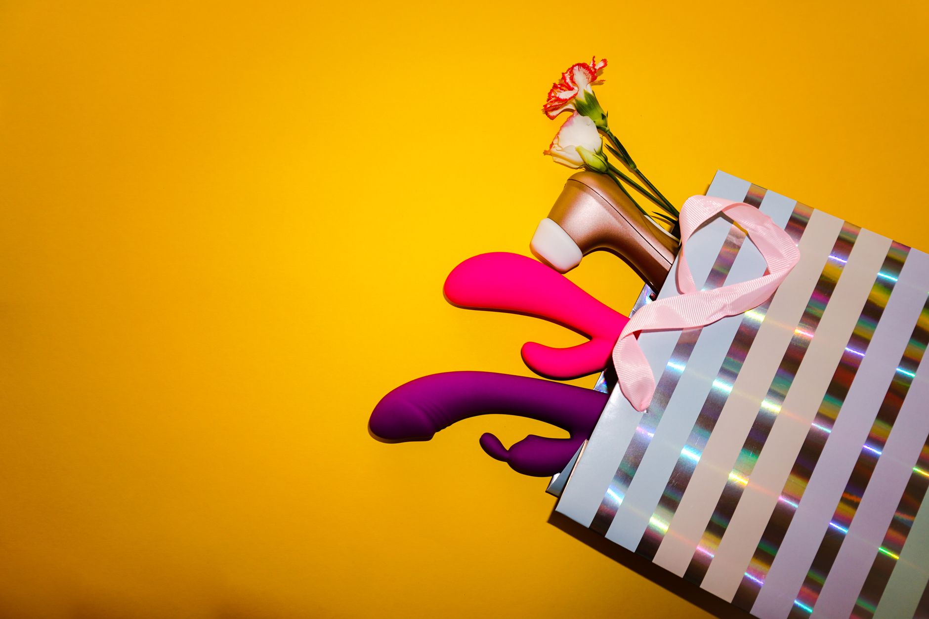 Colorful clitoral vibrators displayed in a holographic gift bag against a bold yellow background